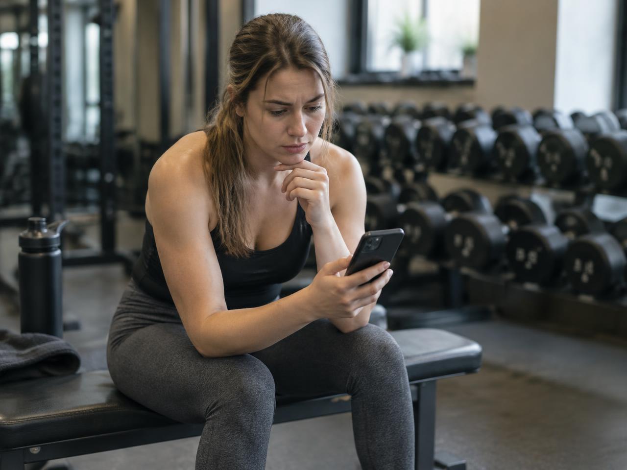 Vrouw zit op een bankje in de sportschool en twijfelt welke oefeningen ze moet doen